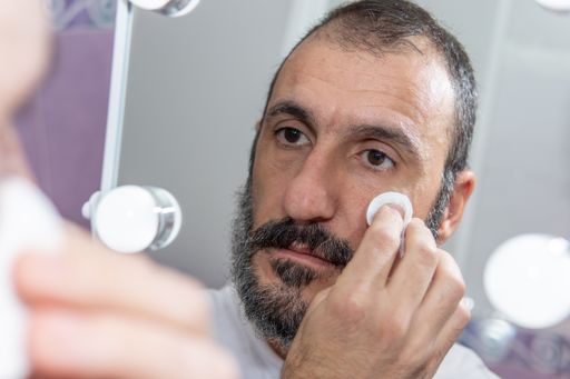 Mature Caucasian bearded man cleaning his face with a cotton pad in front of a mirror, copy space