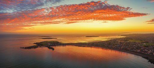A panoramic aerial shot of land surrounded by the sea under an orange sky at sunset