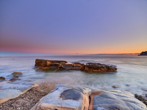 A scenic view of rocks coming out of water under a sunset-colored sky in a long exposure - wallpaper