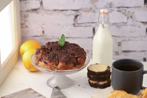 A chocolate cake next to a window accompanied by milk, orange, a cup of coffee, and croissants
