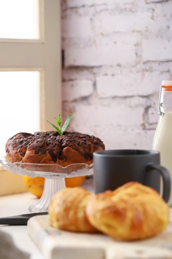 A chocolate cake next to a window accompanied by milk, orange, a cup of coffee, and croissants