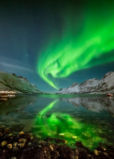 A vertical shot of a green Aurora in the sky over a lake situated in a canyon between snowy hills