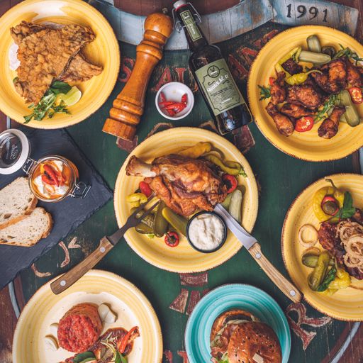 A top view of a set table with baked chicken, pork, a bottle of beer, and a hamburger