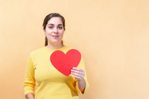 A Portrait of a Caucasian young girl holding a red heart figure on light orange background