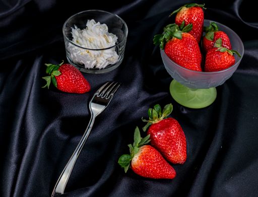 A cup of ripe strawberries with fork and cream on black satin fabric background