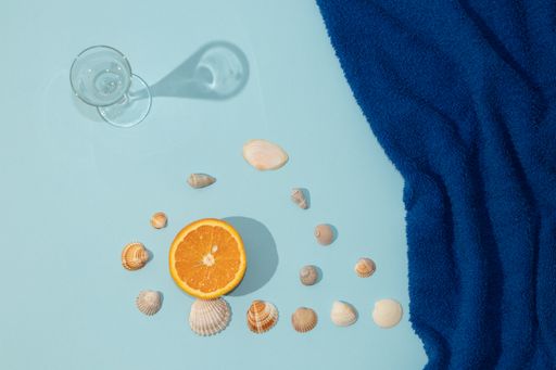 A top view of summer scene with a dark blue beach towel, glass, orange fruit and seashells - Minimal aesthetic