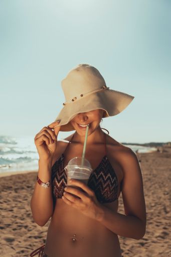 A vertical shot of a young woman in hat smiling and drinking ice coffee near the sea