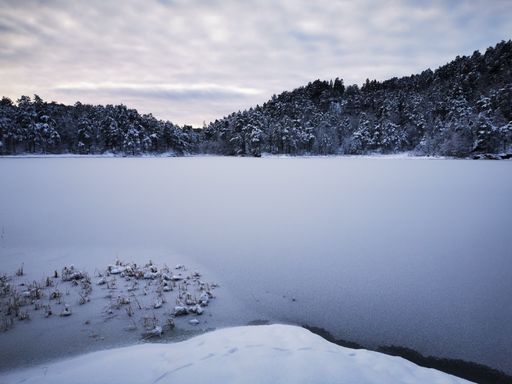 A scenic view of a frozen lake against hills covered with snowy forests in winter