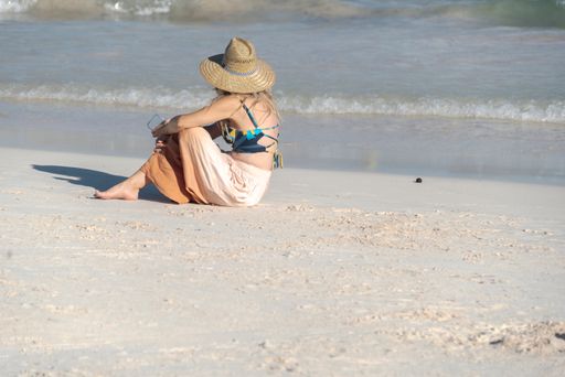 The view of a female wearing a pareo and a hat sitting sidew on the sandy beach looking at the waves