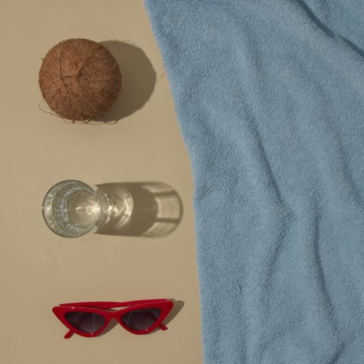 A top view of summer scene with a blue beach towel and coconut, red sunglasses and glass with parasol