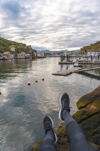 A vertical shot of a person wearing shoes with a view of a coastal town in Pasajes San Pedro, Gipuzkoa, Spain