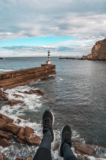 A vertical shot of a person wearing shoes with a view of a beacon at a stone pier in Pasajes San Pedro, Gipuzkoa, Spain