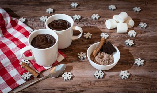 Two cups of chocolate and a bowl of cocoa powder with cinnamon sticks on a wooden table