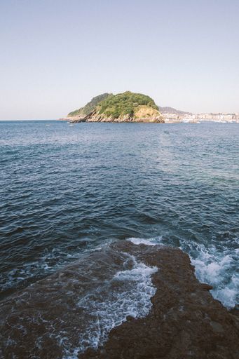 A vertical shot of sea waves hitting the rock with a mountain in the distance under a clear sky