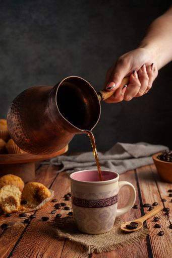A Caucasian hand pouring coffee from long-handled pot into the mug on a wooden table with muffins