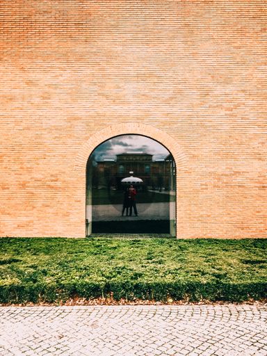 A vertical shot of a brick wall with an arch closed with a glass with two humans reflection on it