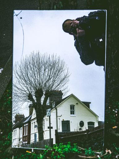 A vertical shot of a male taking a photo of a house near the trees in a mirror