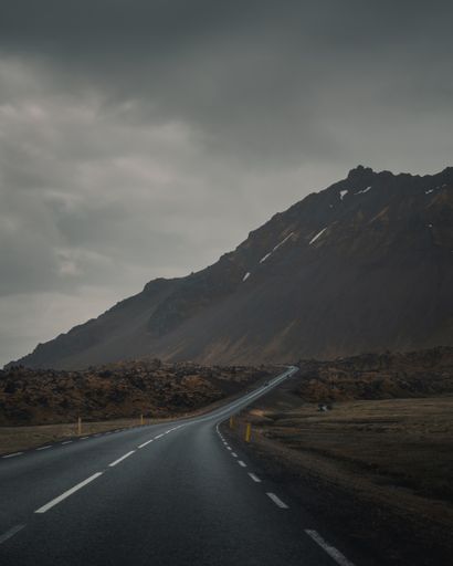 An empty curvy road next to a beautiful rocky mountain under a gray gloomy sky