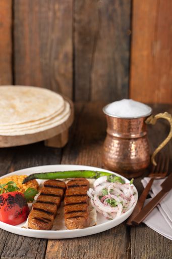 A vertical shot of a plate of kebab and grilled vegetables and a copper jar on a wooden table