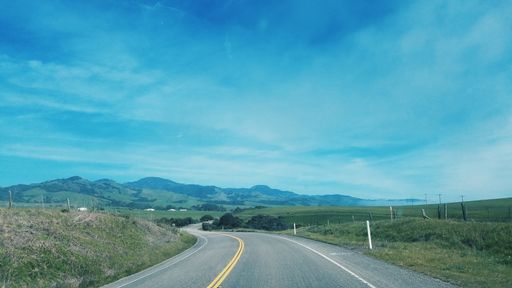 A landscape of a curvy road in a field covered in greenery under a blue cloudy sky in the countryside