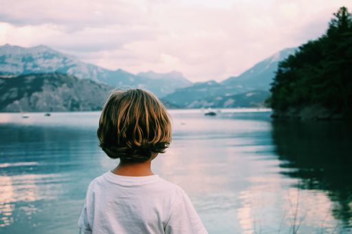 A kid with blonde hair looking at the sea with mountains in the distance shot from behind