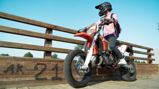 A low angle of a motorcyclist sitting on an orange bike, wearing a helmet against a wooden fence