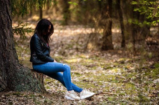 A female wearing jeans and a leather jacket sitting on a tree trunk in the middle of a forest