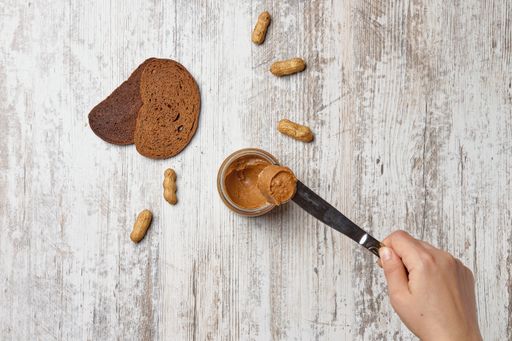 A top view of a hand taking out peanut butter with a knife, bread slices and peanuts on the table