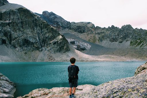 A male with a backpack standing on a cliff enjoying the view of the sea near a mountain shot from behind