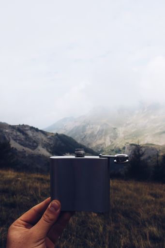 A vertical shot of a person holding a metal flask with a mountain and cloudy sky in the background