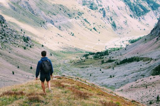 A male with a backpack standing on a cliff enjoying the view surrounded by mountains shot from behind