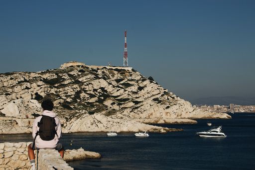 A male with a backpack sitting on a rock fence near the water with a mountain and a clear sky in the background