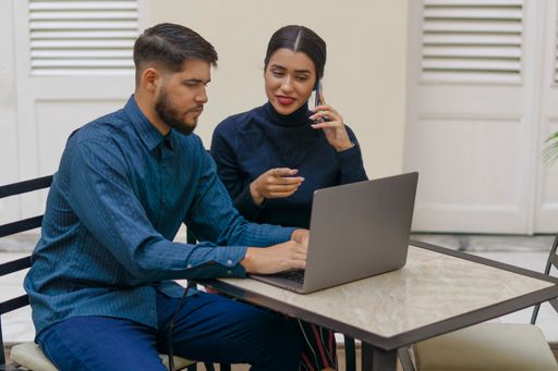 A man and a woman sitting down at a cafe and working on a laptop while talking on the phone