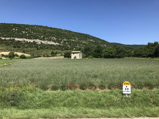 A wide shot of a small cabin in the middle of a grass field surrounded with trees and mountains