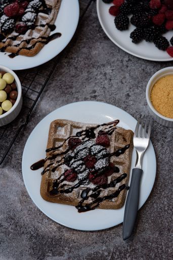 A photo of chocolate waffle with raspberries, a bowl of chocolate, and raspberries plate