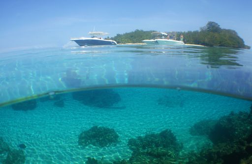 A scenic view of transparent bottom of the sea with boats swimming on the surface against a tropical island