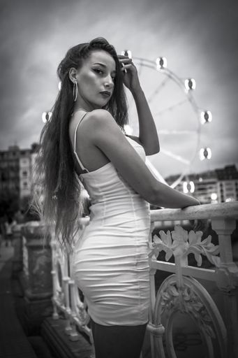 A vertical greyscale shot of an attractive young female posing on a bridge with a Ferris Wheel on the background in San Sebastian