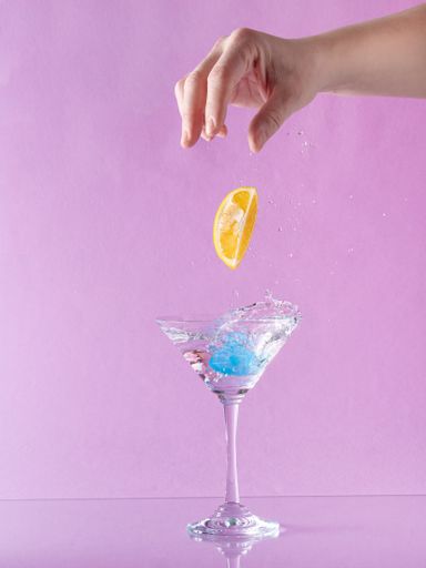 A person throwing lemon into martini glass full of alcohol drink with splashes on pastel pink background