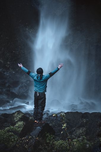 A vertical shot of a man feeling free standing on the rock in front of the waterfall