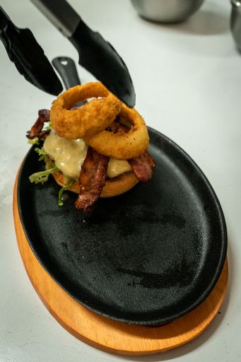 A vertical shot of tongs arranging a burger with bacon and onion rings
