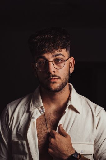 A portrait of a young sexy Hispanic man with glasses and beard wearing a white shirt, black background