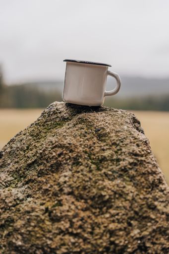 White enamel cup placed on a rock. Close up photo of a metal camping mug on a stone with blurred background.
