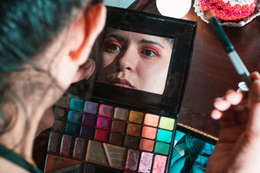 A view of a beautiful Latina girl looking at a small mirror in eyeshadow palette and putting makeup for herself