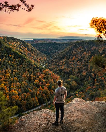 A back view of a man admiring the forest mountains with bright green and orange autumn trees at sunset