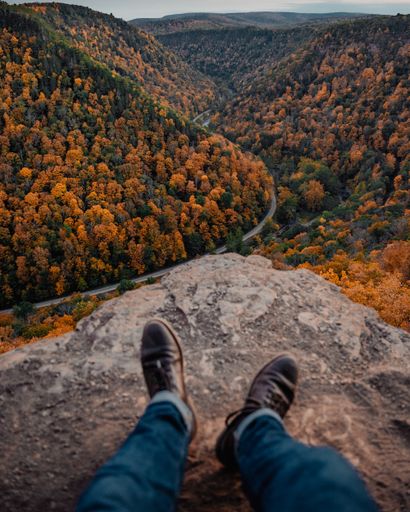 A male feet sitting on the rock against forest mountains with bright green and orange autumn trees