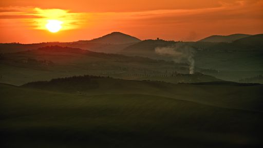 A beautiful panoramic view of a green meadow with white smoke coming out of a rural house chimney at sunset in Tuscany, Italy