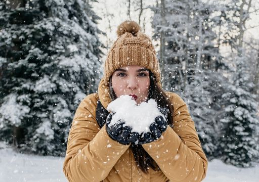 Portrait of beautiful young woman standing outdoor in snowy meadow in woods, blowing snow kiss. Attractive female wearing yellow jacket and knit hat