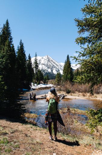 A vertical shot of a female with backpack standing near water and trees with a snowy mountain and clear sky in the background