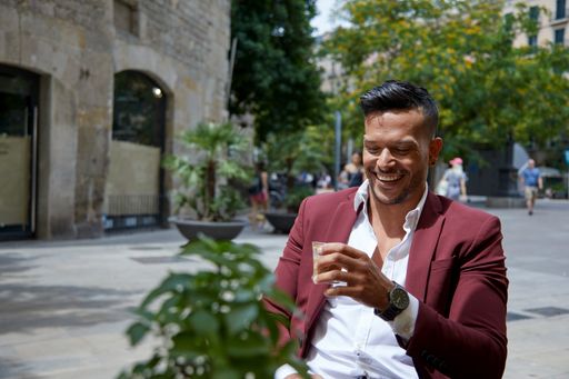 A young Hispanic male in a white shirt and burgundy suit drinking coffee outdoors