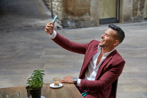 An attractive young Hispanic male in red suit taking a selfie on terrace with coffee